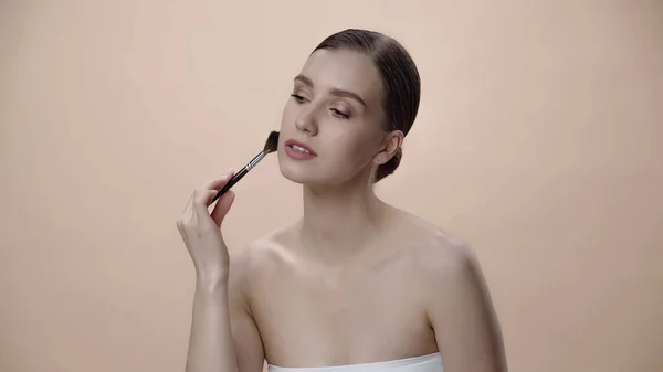 Pretty young woman with bare shoulders applying face powder while holding cosmetic brush isolated on beige — Stockfoto