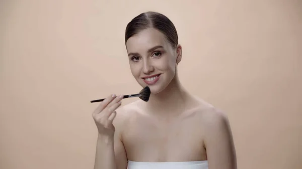 Mujer joven feliz con hombros desnudos aplicando polvo facial mientras sostiene cepillo cosmético aislado en beige - foto de stock