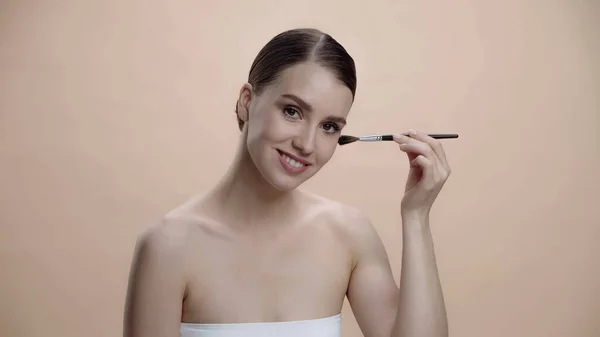 Positive young woman with bare shoulders applying face powder while holding cosmetic brush isolated on beige — Photo de stock