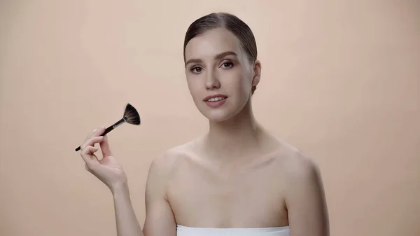 Young woman with bare shoulders holding cosmetic brush isolated on beige — Fotografia de Stock