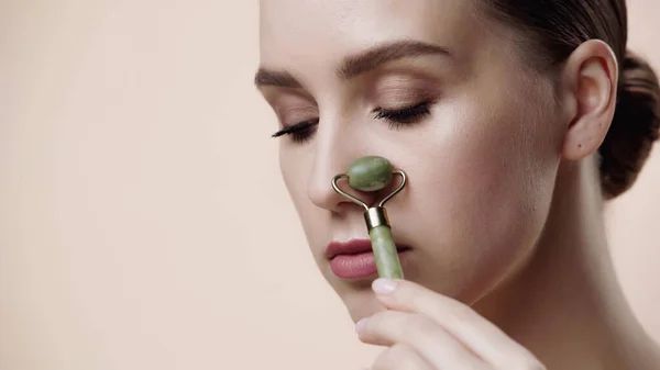 Young woman using jade roller on nose isolated on beige — Stockfoto