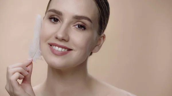 Alegre joven sosteniendo pluma blanca cerca de la cara aislada en beige - foto de stock