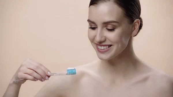 Happy woman with bare shoulders holding toothbrush with toothpaste isolated on beige — Stockfoto