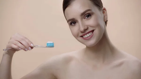 Cheerful woman with bare shoulders holding toothbrush with toothpaste isolated on beige — Photo de stock
