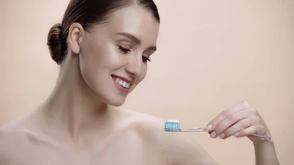Joyful woman with bare shoulders holding toothbrush with toothpaste isolated on beige — Stockfoto