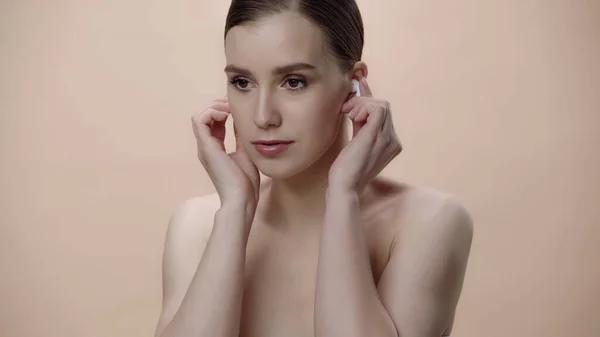 Young woman with bare shoulders adjusting earphones isolated on beige — Stockfoto