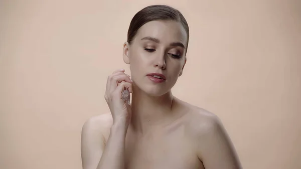 Young woman with bare shoulders holding bottle and applying luxurious perfume isolated on beige — Fotografia de Stock