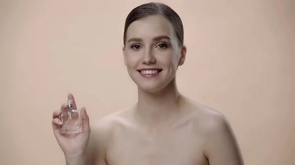 Щаслива молода жінка з голими плечима тримає пляшку з розкішними парфумами ізольована на бежевому — стокове фото