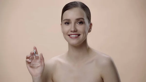 Happy young woman with bare shoulders holding bottle with luxurious perfume isolated on beige — стоковое фото