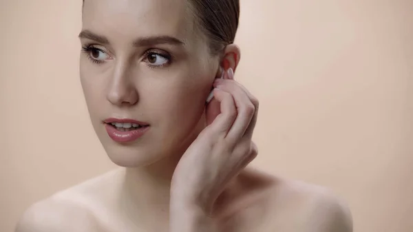 Young woman with bare shoulders adjusting earphone isolated on beige — Stock Photo