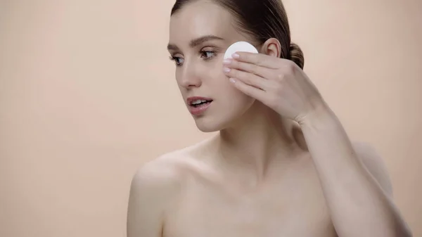 Young woman with bare shoulders holding cotton pad near face isolated on beige — Foto stock