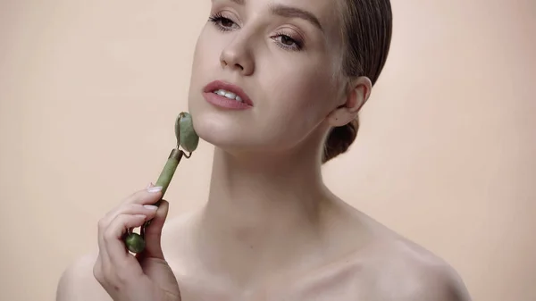 Young woman with bare shoulders massaging face with jade roller isolated on beige — Photo de stock