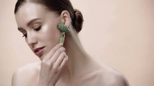 Pretty woman with bare shoulders massaging face with jade roller isolated on beige — Stock Photo