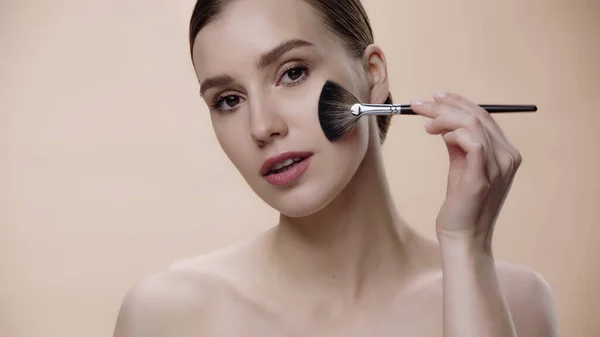 Young woman with bare shoulders applying face powder on cheek isolated on beige - foto de stock