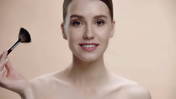 Joyful woman with bare shoulders holding cosmetic brush isolated on beige — Photo de stock