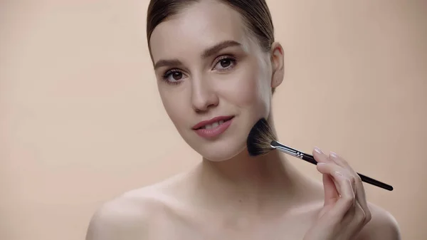 Mujer alegre con hombros desnudos aplicando polvo facial mientras sostiene cepillo cosmético aislado en beige — Stock Photo