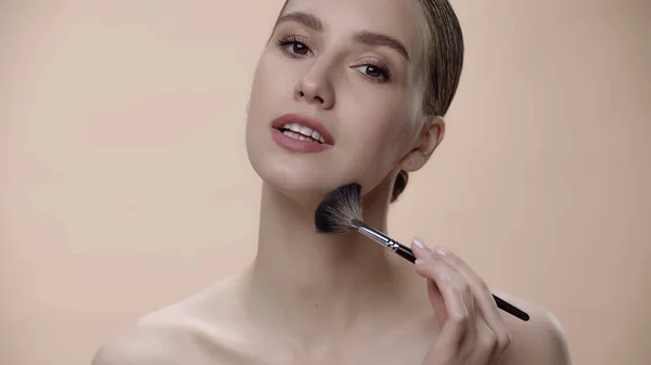 Mujer joven aplicando polvo facial con cepillo cosmético aislado en beige - foto de stock