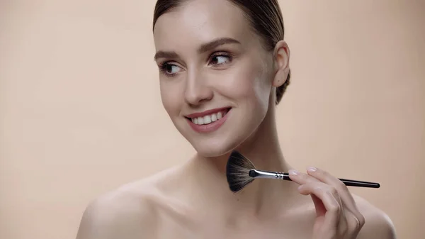Pleased woman with bare shoulders applying face powder while holding cosmetic brush isolated on beige - foto de stock