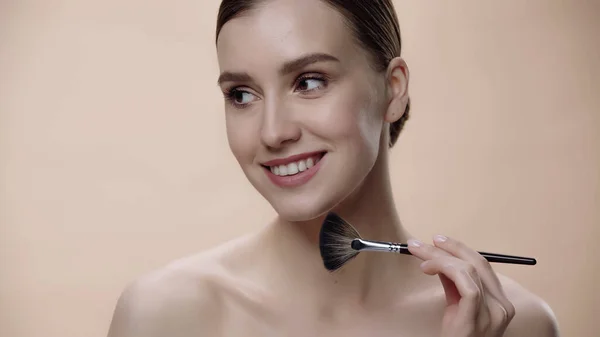 Pleased woman with bare shoulders applying face powder while holding cosmetic brush isolated on beige — Fotografia de Stock