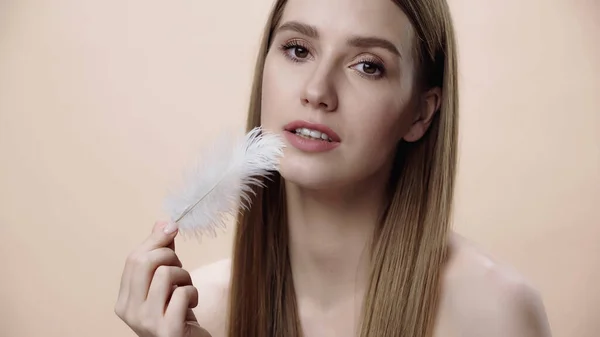 Young woman with bare shoulders touching face with feather isolated on beige — стоковое фото