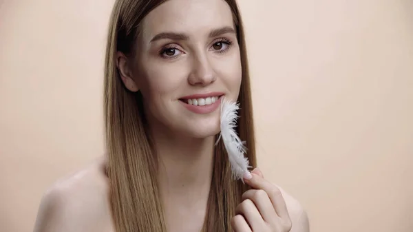 Happy young woman with bare shoulders touching face with white feather isolated on beige — Foto stock