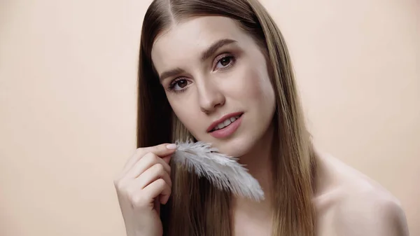 Pretty woman with bare shoulders touching face with white feather isolated on beige — Foto stock