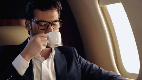 Businessman in eyeglasses drinking coffee in private plane — Stock Photo