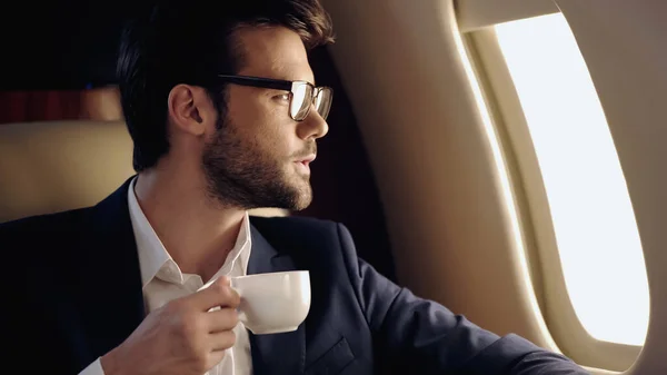 Homme d'affaires barbu en lunettes tenant une tasse en jet privé — Photo de stock