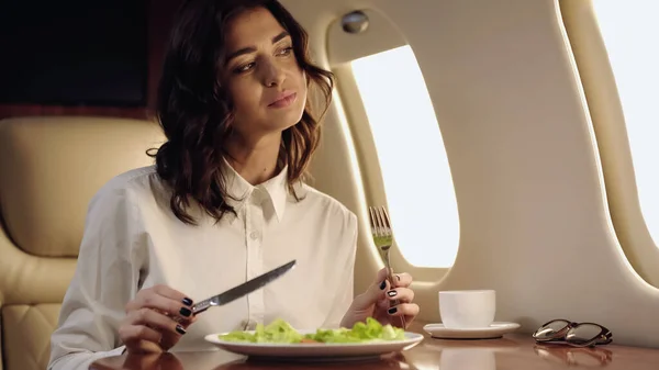 Приємна бізнес-леді тримає столові прибори біля свіжого салату і чашки в приватному реактивному літаку — стокове фото