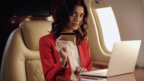 Joven mujer de negocios con tarjeta de crédito y mirando a la cámara cerca de la computadora portátil en jet privado - foto de stock