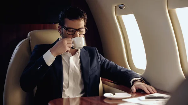Empresario en anteojos bebiendo café cerca de smartphone y papeles en avión privado - foto de stock