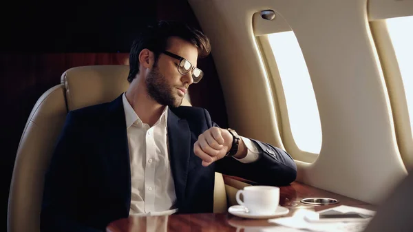 Businessman looking at wristwatch near coffee and cellphone in private plane — Stock Photo