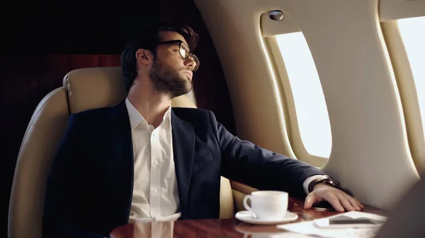 Businessman looking at window near coffee and smartphone in private jet — Stock Photo