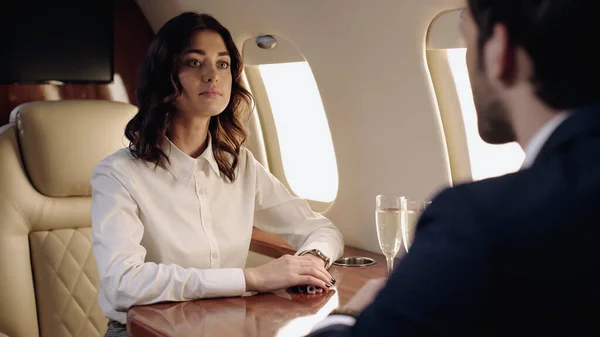 Бізнес-леді дивиться на розмитого хлопця біля келихів шампанського в приватному літаку — стокове фото