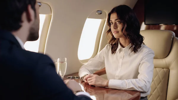 Young businesswoman looking at blurred boyfriend in formal wear near champagne in private plane — Stock Photo