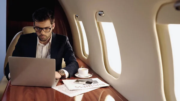 Empresario usando portátil cerca de café y teléfono inteligente en jet privado - foto de stock