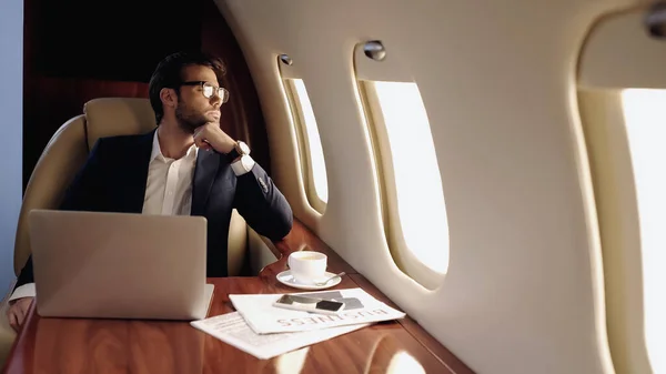 Businessman looking at window near devices, coffee and newspapers in private jet - foto de stock