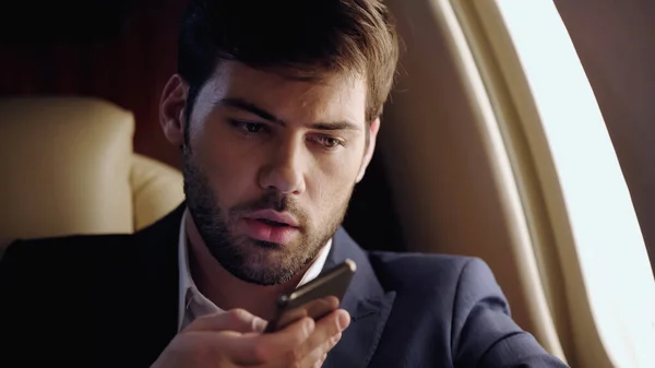 Bearded businessman recording voice message on blurred smartphone in private jet — стоковое фото