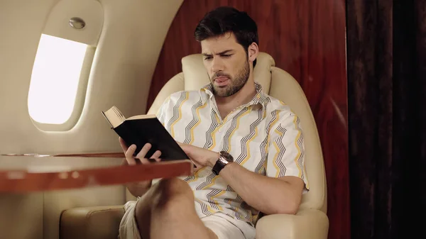 Barbudo hombre sobresaliendo lengua mientras lee libro en avión privado - foto de stock