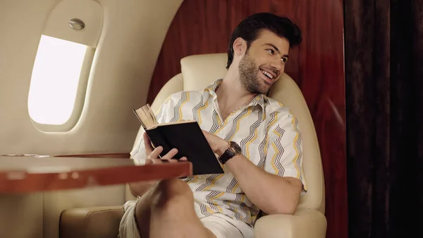 Smiling man holding book and looking away in private jet — Stockfoto