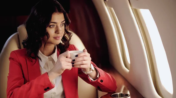 Businesswoman holding cup looking at window in private plane — Foto stock