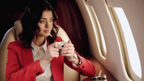 Businesswoman holding cup looking at window in private plane — Stock Photo