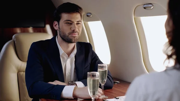 Smiling businessman looking at girlfriend near glasses of champagne in private plane — Stock Photo