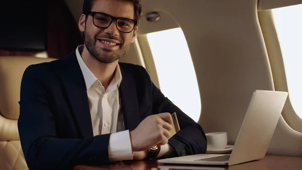 Smiling businessman holding credit card and looking at camera near laptop and cup in private jet — Fotografia de Stock