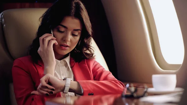 Businesswoman talking on smartphone and looking at wristwatch near cup in private plane — Stock Photo