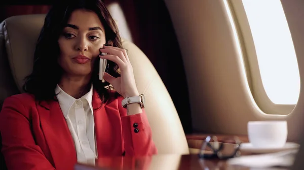 Businesswoman talking on smartphone near blurred cup in private plane — Stock Photo