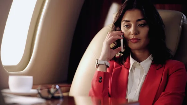 Businesswoman talking on cellphone near blurred cup and eyeglasses in private plane — Stock Photo