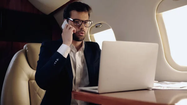Empresario hablando en smartphone cerca de laptop y papeles en avión privado - foto de stock