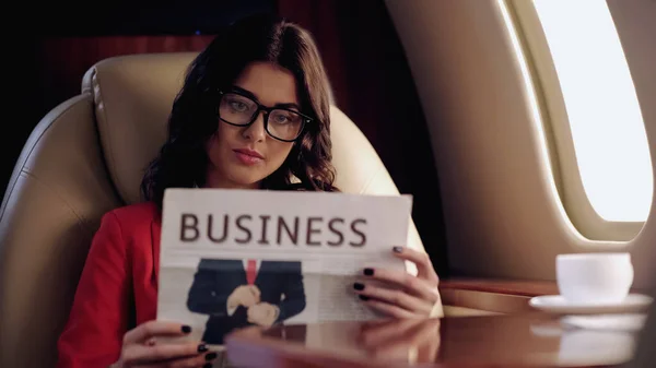 Businesswoman in eyeglasses reading newspaper near coffee in private plane — Stock Photo