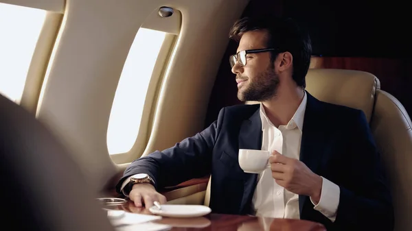 Smiling businessman holding cup and looking at window in private plane — Stock Photo